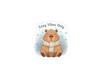 May include: A sticker featuring a cartoon capybara with a blue and white striped scarf, surrounded by snowflakes. The text "Cozy Vibes Only" is above the capybara.