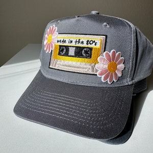 以下が含まれることがあります： 「made in the 80's」と書かれたレトロなカセットテープのパッチが付いたグレーの野球帽。 ピンクと黄色のデイジーパッチが2つ飾られています。 帽子はカーブしたつばと構造化されたクラウンを備えています。