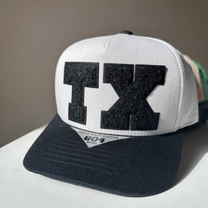 Puede incluir: Gorra de béisbol blanca y negra con un parche de fieltro negro que dice "TX" en la parte delantera. La gorra tiene una visera negra y una parte trasera de malla blanca. La etiqueta de la gorra dice "Premium Quality Headwear 804 Kamel"