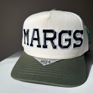 May include: A white and green baseball cap with the word "MARGS" embroidered in black on the front. The cap has a black and white label on the back that says "PREMIUM QUALITY HEADWEAR 804 KAMEL".