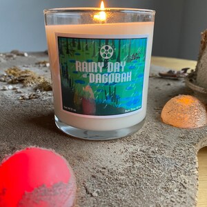 Op de afbeelding: Een brandende kaars in een heldere glazen pot met het label "Rainy Day at Dagobah". De kaars staat op een zanderige ondergrond met decoratieve elementen. De brandtijd van de kaars is 40 uur.