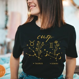 May include: Black t-shirt with a gold graphic of the Taurus and Gemini zodiac signs with the word "cusp" above them. The graphic also includes flowers and stars.