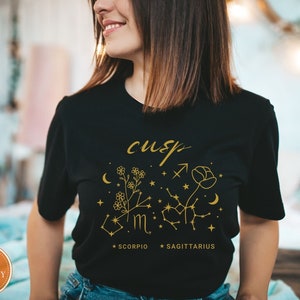 May include: Black t-shirt with a gold graphic design featuring the word "cusp", floral elements, constellations, and the words "Scorpio" and "Sagittarius". The shirt has a round neckline.