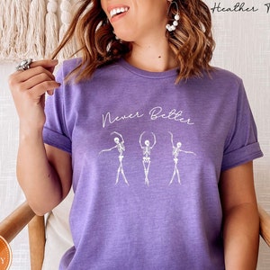 May include: A purple t-shirt with the text "Never Better" and a graphic of three skeletons dancing.