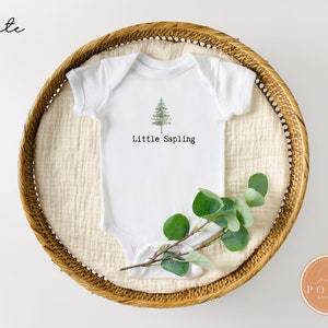 Könnte beinhalten: Weißer Baby-Body mit einem grünen Baum-Motiv und dem Schriftzug "Little Sapling". Der Body liegt in einem geflochtenen Korb, zusammen mit einem Zweig grüner Blätter.