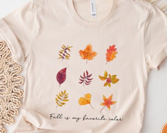 May include: Cream-colored t-shirt featuring a watercolor design of nine colorful autumn leaves. The text "Fall is my favorite color" is printed below the leaves. The shirt has a round neckline and short sleeves.