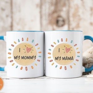 May include: Two white ceramic mugs with blue handles. The mugs have a watercolor-style sun design with the text "I love my mommy" and "I love my mama" on each mug.