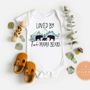 May include: White baby onesie with a watercolor mountain scene and two black bears. The text "Loved by Two Mama Bears" is printed on the onesie.