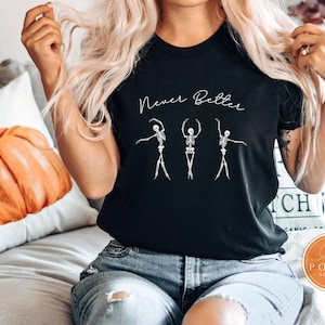 May include: Black t-shirt with the text "Never Better" above a graphic of three skeleton ballerinas in ballet poses. The shirt is a crew neck and is made of a soft material. The image also shows a pumpkin pillow.