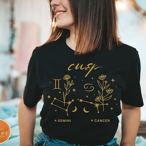 May include: Black t-shirt with a gold graphic of the Gemini and Cancer zodiac signs with the word "cusp" above them. The graphic also includes flowers and stars.