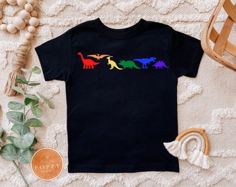 May include: Black t-shirt with a rainbow dinosaur design. The dinosaurs are in a line and include a stegosaurus, a pterodactyl, a triceratops, a brachiosaurus, and a tyrannosaurus rex.