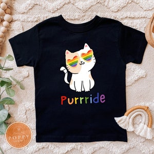 May include: Black t-shirt featuring a cartoon cat wearing rainbow heart-shaped sunglasses. The cat is white and tan, with the word "Purrrride" in rainbow colors below the cat. The shirt is designed for children.
