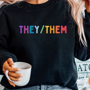 May include: A black sweatshirt with the text "THEY/THEM" in rainbow colors.