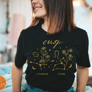May include: Black t-shirt with gold text and graphics. The design includes the word "cusp", floral illustrations, and the Cancer and Leo zodiac signs. The shirt is a crew neck style.