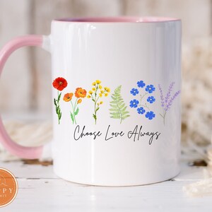 May include: White ceramic mug with a pink handle and interior. The mug features a floral design with red, orange, yellow, green, blue, and purple flowers. The words "Choose Love Always" are written in black script.