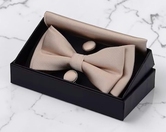 May include: A formal beige bow tie, pocket square, and cufflink set presented in a black box. The bow tie and pocket square have a textured appearance, and the cufflinks are round. The set is displayed on a marble surface.