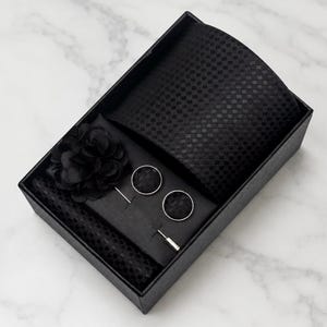 May include: A black gift box containing a black necktie with a subtle checkered pattern, a black fabric flower lapel pin, a pair of round black cufflinks with a matching checkered texture, and a black pocket square with a fine mesh pattern.