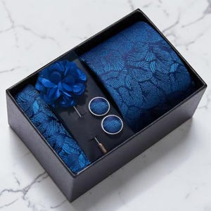 Peut inclure: Un ensemble de cravate bleu dans une boîte noire. L'ensemble comprend une cravate, une pochette, une fleur de revers et des boutons de manchette. La cravate et la pochette ont un motif de feuilles. La fleur de revers est bleu foncé.