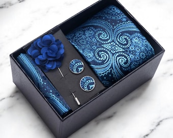 May include: A navy blue gift box containing a paisley patterned tie, pocket square, lapel flower, and cufflinks. The set includes a tie, pocket square, lapel flower, and cufflinks. The accessories are in shades of blue.