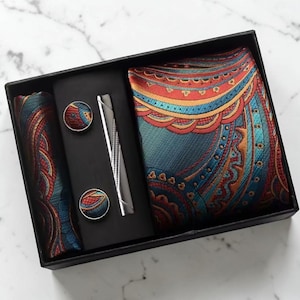Peut inclure: Un coffret cadeau contenant une cravate, une pochette et des boutons de manchette à motifs cachemire bleu sarcelle, orange et bleu. L'ensemble comprend une pince à cravate argentée et deux boutons de manchette ronds assortis, dans une boîte noire.