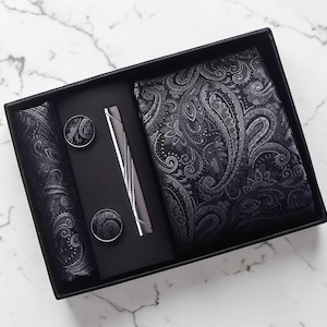 May include: A black gift set featuring a paisley patterned tie, pocket square, cufflinks, and a silver pen. The tie and pocket square display a detailed silver design on a black backdrop. The cufflinks are round with matching paisley patterns.
