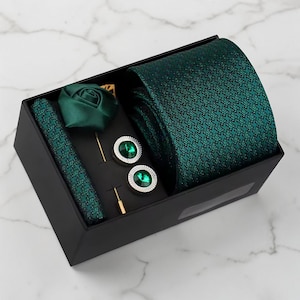 May include: A dark green tie set in a black box. The set includes a tie, pocket square, lapel pin, and cufflinks. The tie and pocket square have a small, repeating pattern. The cufflinks have a green stone center.
