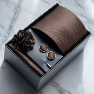 May include: A brown tie set presented in a black box. The set includes a tie, pocket square, cufflinks, lapel flower, and tie pin. The tie and pocket square have a ribbed texture. The cufflinks and lapel flower are a darker shade of brown.