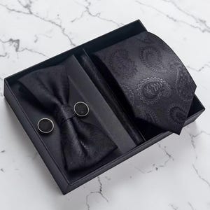 May include: A black formal accessory set in a gift box. The set includes a paisley-patterned tie, bow tie, pocket square, and cufflinks. The items are black with a subtle pattern, presented in a black box.