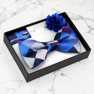 May include: A formal accessory set presented in a black box. The set includes a blue patterned bow tie, a matching pocket square, and a blue lapel flower. The bow tie and pocket square feature a geometric design in shades of blue, purple, and white.