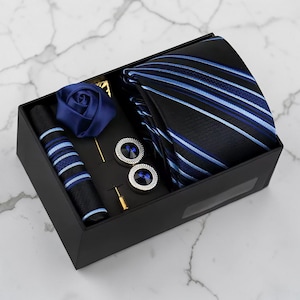 May include: A black gift box containing a striped tie, pocket square, cufflinks, and a lapel flower. The tie features blue and white stripes. The cufflinks have a blue centre. The lapel flower is a deep blue rose.