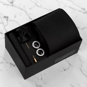 May include: A black gift box containing a black necktie with a herringbone pattern, a matching pocket square, two round silver-toned cufflinks with black centres, and a gold-toned lapel pin with a small black accent piece.