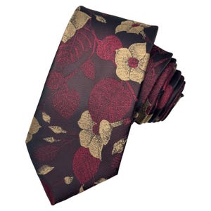 Puede incluir: Una corbata enrollada con un estampado floral. La corbata presenta un fondo burdeos oscuro con diseños florales dorados y burdeos. La corbata está hecha de un material liso, posiblemente seda.