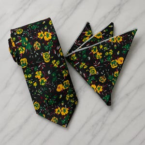 Può includere: Una cravatta nera e una pochette da taschino piegata abbinata, entrambe caratterizzate da un vivace motivo floreale con fiori gialli e verdi, insieme a piccoli accenti bianchi e rosso scuro sparsi sul tessuto scuro di entrambi gli articoli.