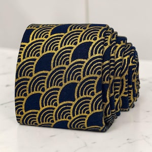 Könnte beinhalten: Eine aufgerollte Krawatte mit dunkelblauem Stoffgrund und einem sich wiederholenden goldfarbenen Metallic-Muster aus konzentrischen Halbkreisen. Der Stoff hat eine strukturierte Optik und die goldenen Linien bilden ein geometrisches Wellenmuster.