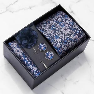 May include: A navy blue floral patterned tie set in a black box. The set includes a tie, pocket square, lapel flower, and cufflinks. The floral pattern features blue, white, and gray flowers. The cufflinks and lapel flower are solid navy blue.