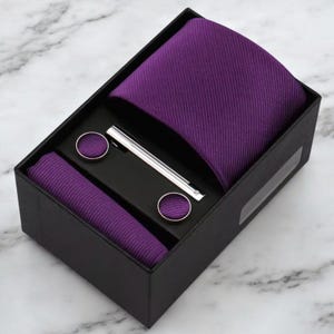 May include: A purple tie, pocket square, cufflinks, and tie clip set in a black box. The tie and pocket square have a ribbed texture. The cufflinks are round with a purple center. The tie clip is silver.