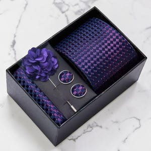May include: A boxed set containing a navy and purple chequered tie, pocket square, and cufflinks. A purple flower lapel pin and tie bar are also included. The set is presented in a black box.