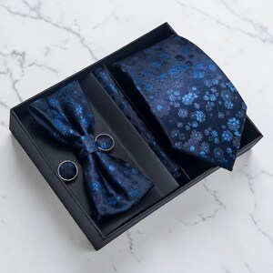 Peut inclure: Un ensemble d'accessoires formels bleu marine dans une boîte noire. L'ensemble comprend une cravate, un nœud papillon, une pochette et des boutons de manchette. Chaque article présente un motif floral dans les tons de bleu. La boîte est sur une surface en marbre blanc.