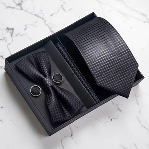 May include: A black formal wear set in a gift box. The set includes a tie, bow tie, pocket square, and cufflinks. All items have a checkered pattern. The box is black and sits on a white marble surface.
