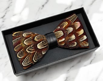 May include: A brown and beige feather bow tie with a black leather strap. The bow tie is in a black box.