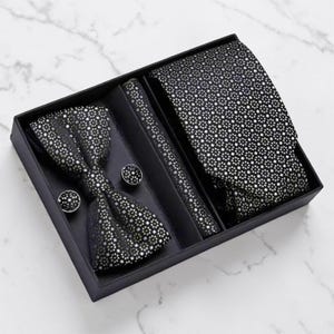 May include: A black gift box containing a bow tie, tie, pocket square, and cufflinks. The bow tie, tie, and pocket square have a repeating silver dot pattern. The cufflinks are round with a matching dot pattern.