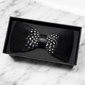 May include: A black and white feather bow tie with a black leather strap. The bow tie is in a black box.