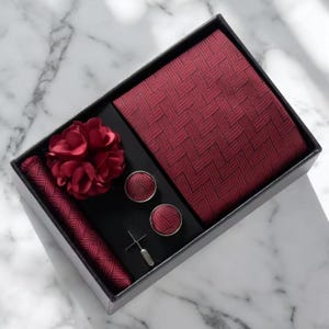 May include: A burgundy tie set presented in a black box. The set includes a tie, pocket square, cufflinks, lapel flower, and tie pin. The tie and pocket square feature a woven design. The cufflinks and tie pin are silver.