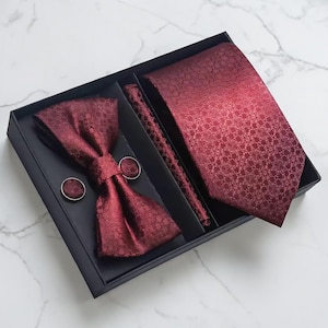 May include: A burgundy formal wear set in a black box. The set includes a patterned tie, bow tie, pocket square, and cufflinks. The items have a repeating floral pattern. The box is on a white marble surface.