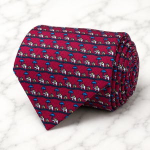 May include: A rolled necktie in a deep magenta red color featuring a repeating pattern of small white elephants holding bright blue umbrellas. The fabric has a fine diagonal ribbed texture, and the elephants are arranged in horizontal rows.