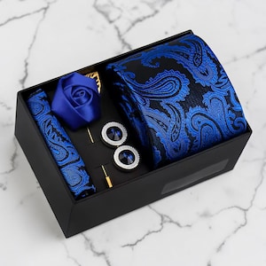 Peut inclure: Un coffret noir contenant une cravate, une pochette, des boutons de manchette et une fleur de revers bleus à motifs cachemire. La cravate et la pochette présentent un motif cachemire bleu détaillé sur fond noir. Les boutons de manchette sont argentés avec un centre bleu.
