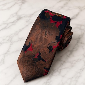 May include: A necktie featuring a brown base with a woven floral pattern in shades of dark navy blue and vibrant red. The fabric has a subtle sheen and a textured appearance, with the tie folded over itself to show the pointed end and the layered construction.
