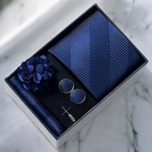 May include: A navy blue men's accessory set in a gift box. The set includes a tie, pocket square, cufflinks, lapel flower, and tie pin. The tie and pocket square have a dotted pattern. The cufflinks and lapel flower are solid navy blue.