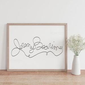 Pode incluir: Uma impressão emoldurada com o nome "Jeremy Bearimy" em um desenho de linha contínua. A obra de arte está em uma moldura de madeira clara e está em uma superfície de madeira. Um vaso branco com flores brancas está à direita da moldura.
