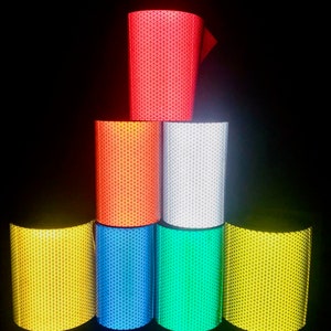 May include: A stack of six rolls of reflective tape in different colors: red, orange, white, blue, green, and yellow. The tape has a diamond pattern.